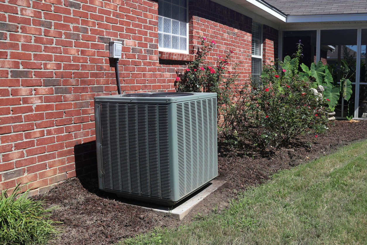 A green-grey central AC unit is installed outside a brick home. 