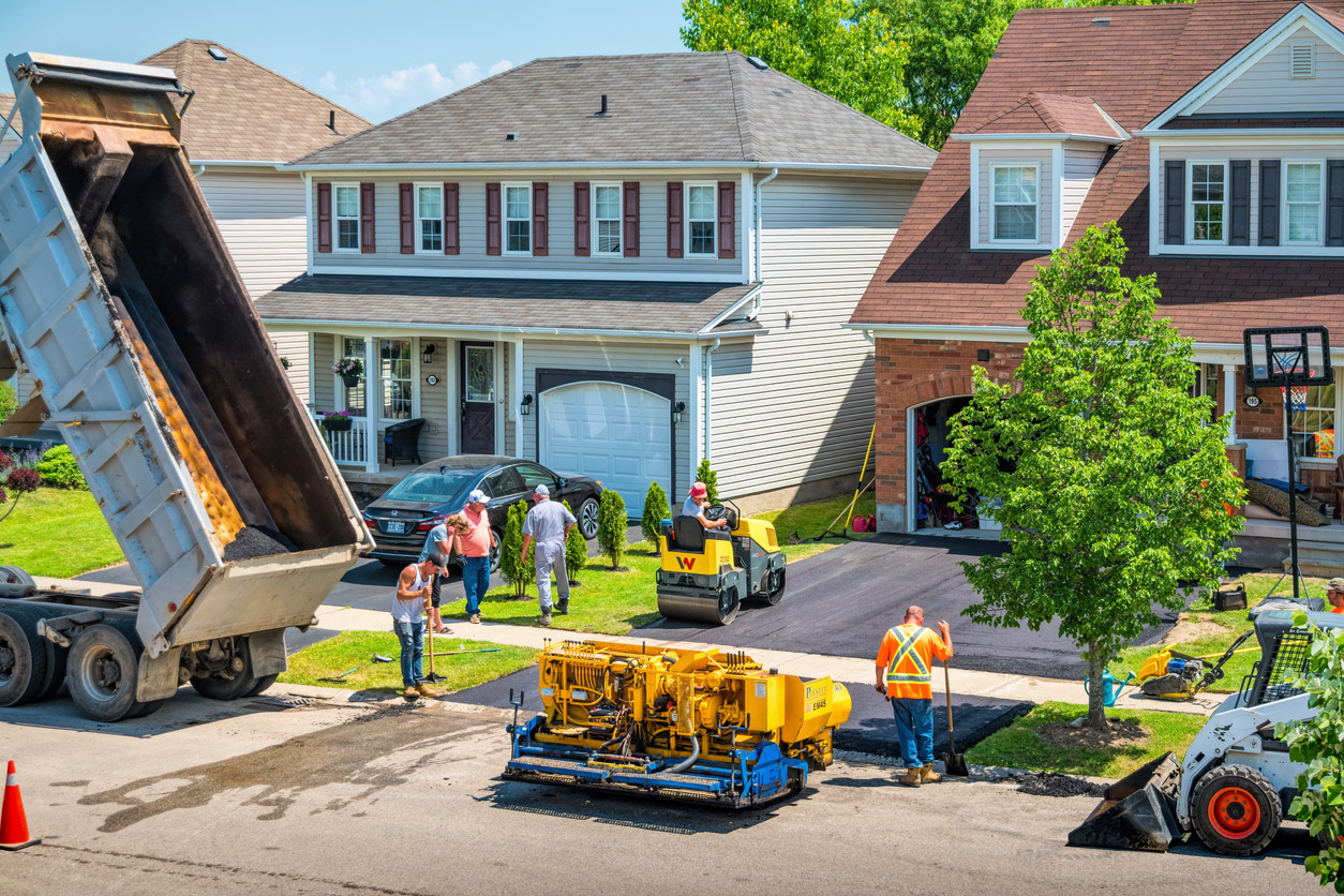 A construction crew paves a driveway with asphalt in front of a two-story house in a residential neighborhood.