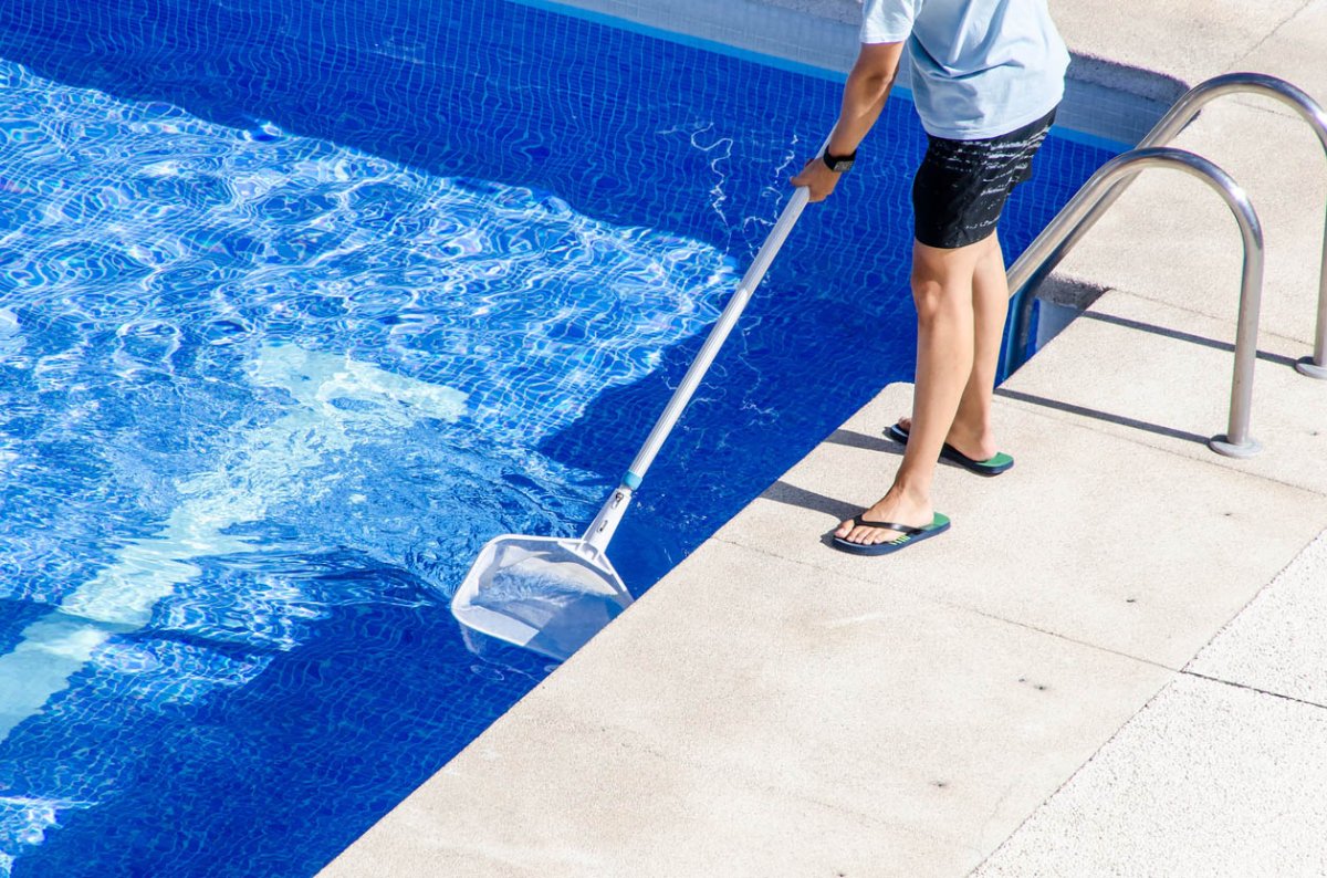 A person with only their legs visible skims a swimming pool to remove debris.