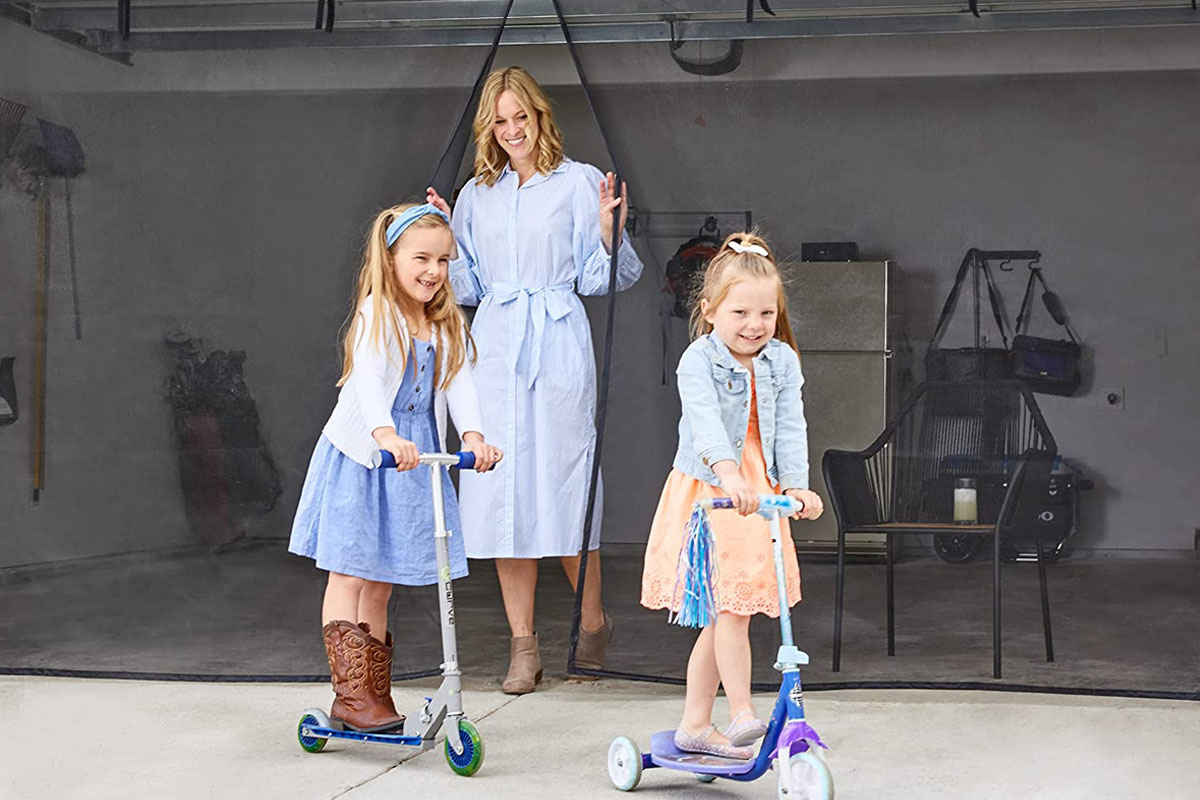 A woman peeks through the closure on the best garage door screens option to smile out at two young girls riding scooters on the driveway