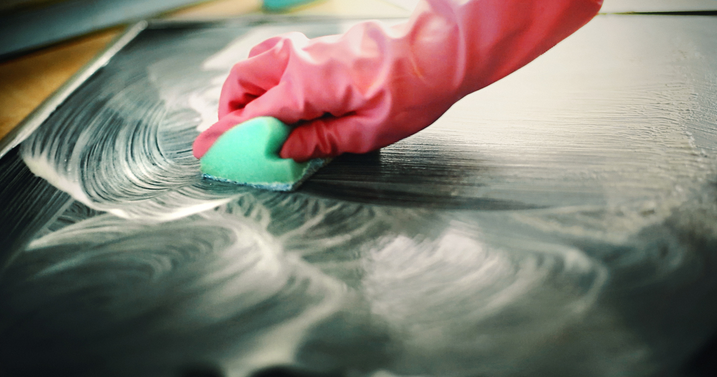 Closeup of unrecognizable person scrubbing the surface of cooking stove with a spone and some detergent. The person is wearing pink protective gloves. Side view, low angle shot.