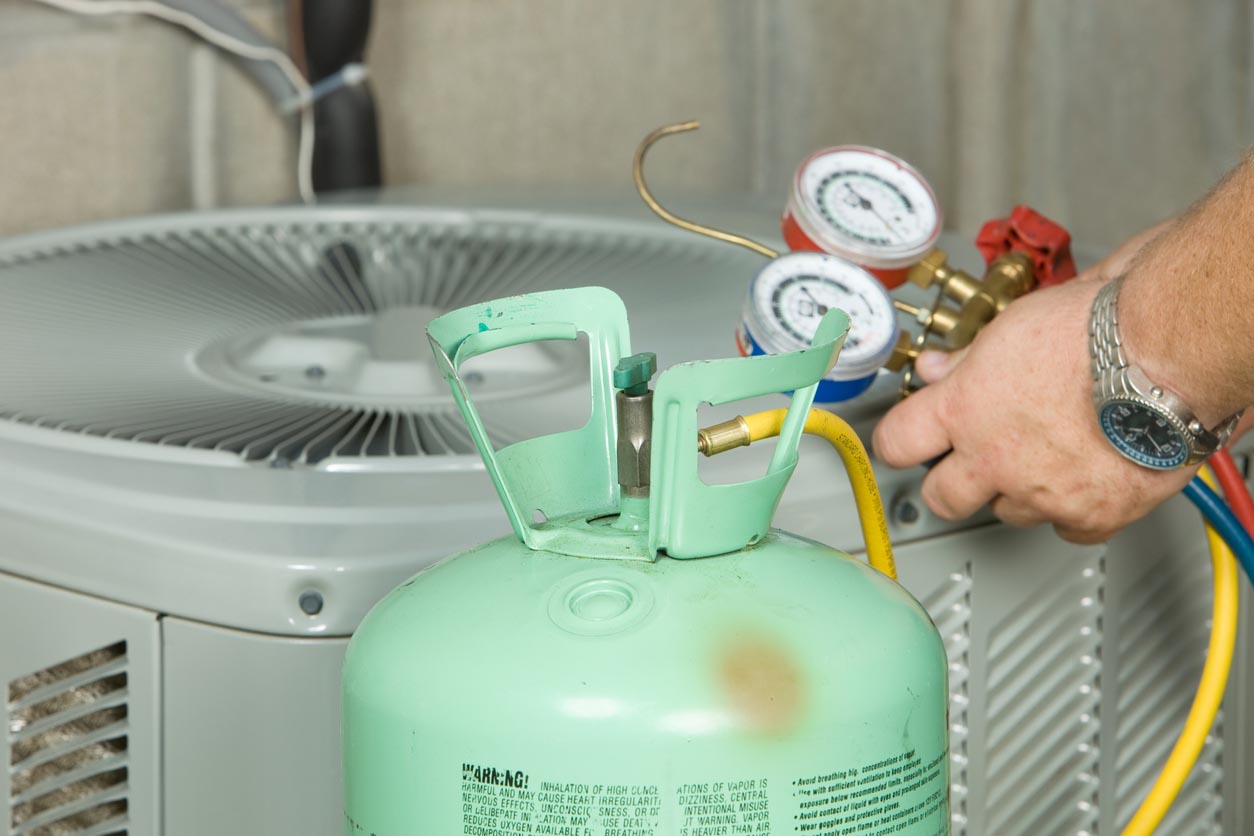 A worker with only their hands visible inspects the coolant level of an outside air conditioning unit.