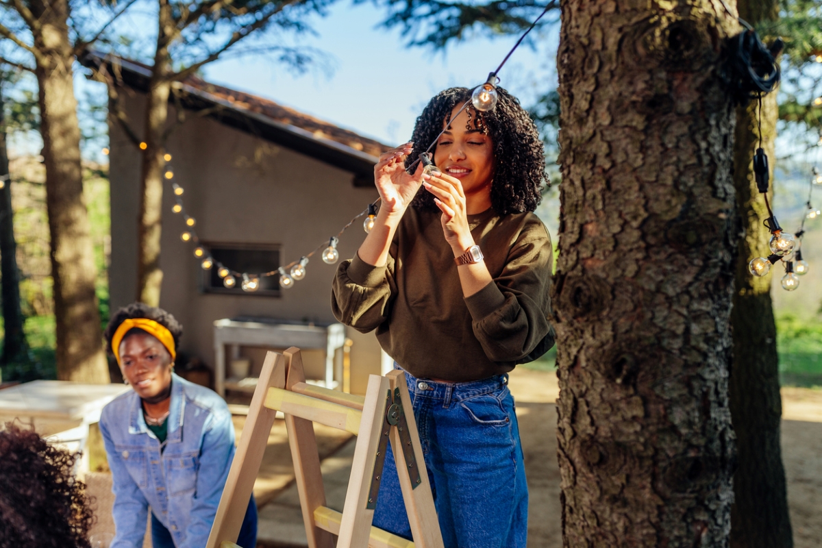 Two women hanging string lights from tree from ladder