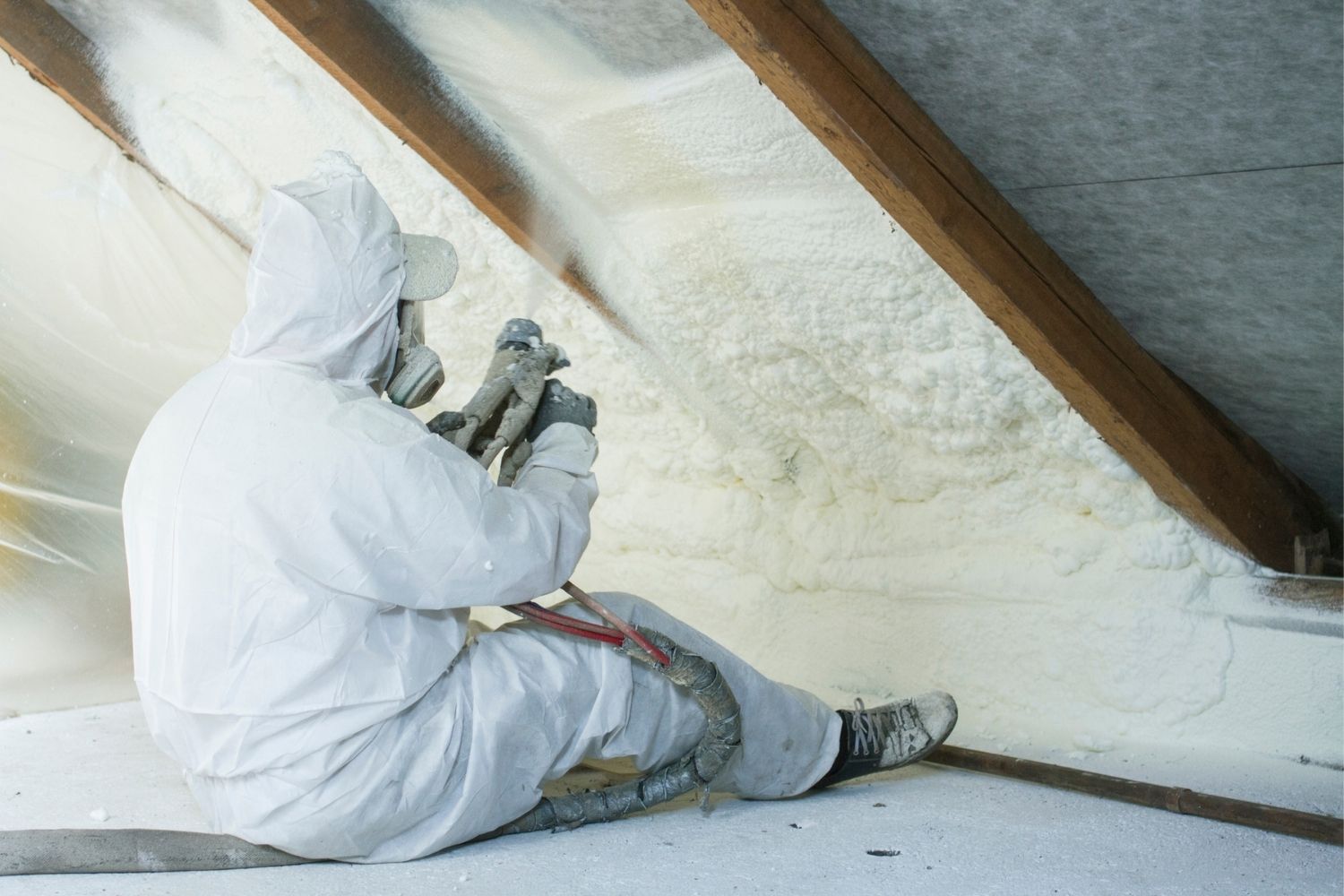 A worker wears personal protective equipment and sprays foam insulation underneath a roof in an attic.