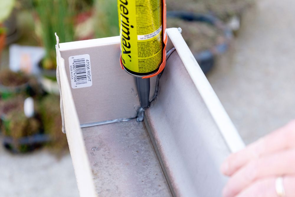 Person squeeze a gray caulk sealant onto the seams of a white gutter