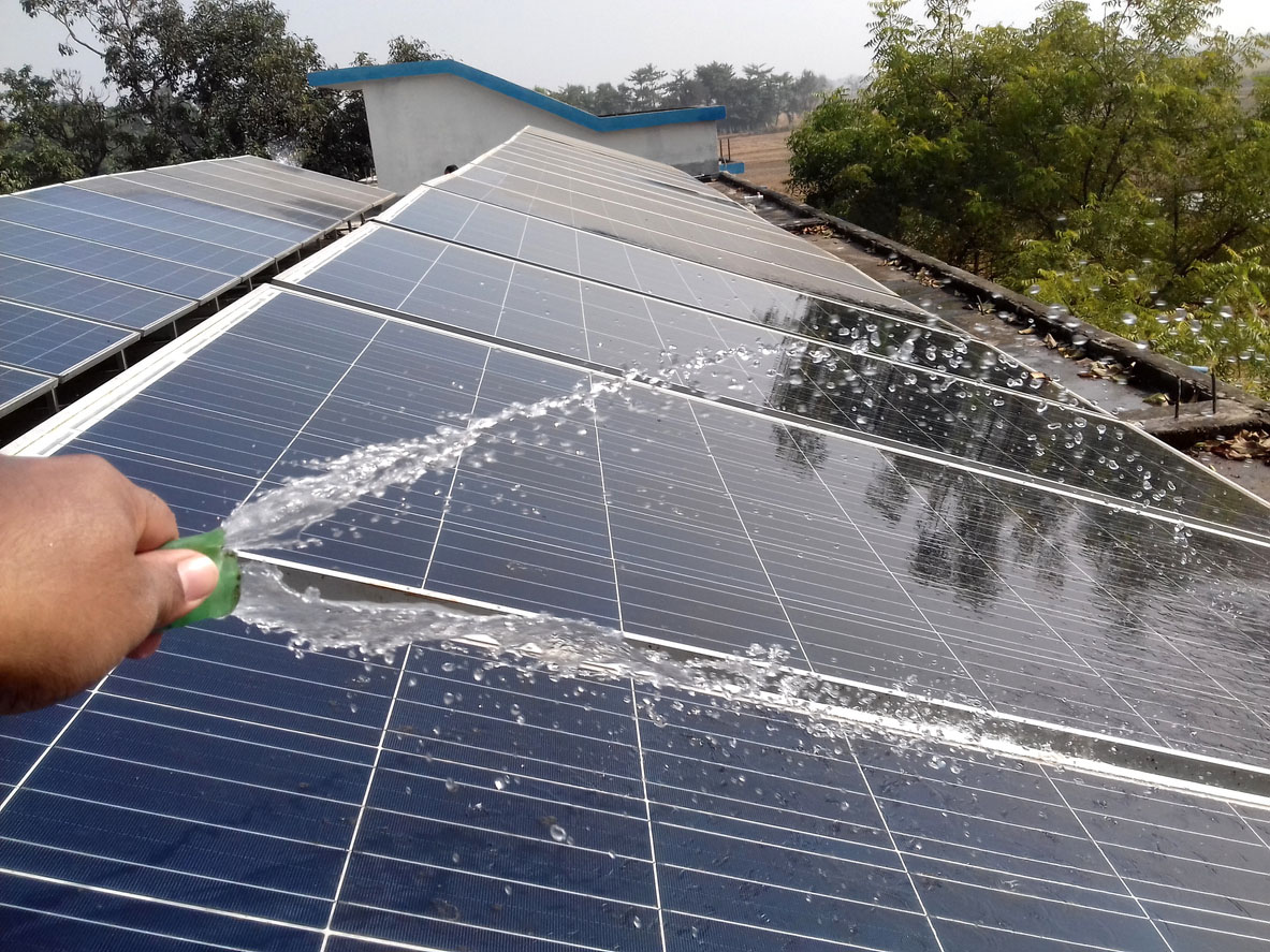 A person with only their hand visible cleans roof solar panels by spraying water from a hose.