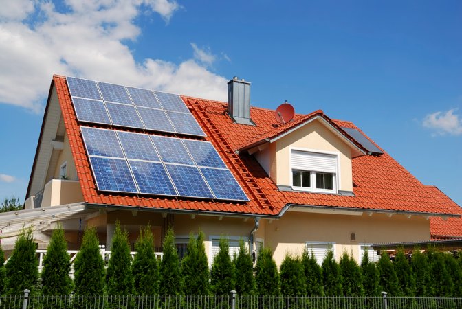 A house with a red roof topped with solar panels against a blue sky with minimal clouds.