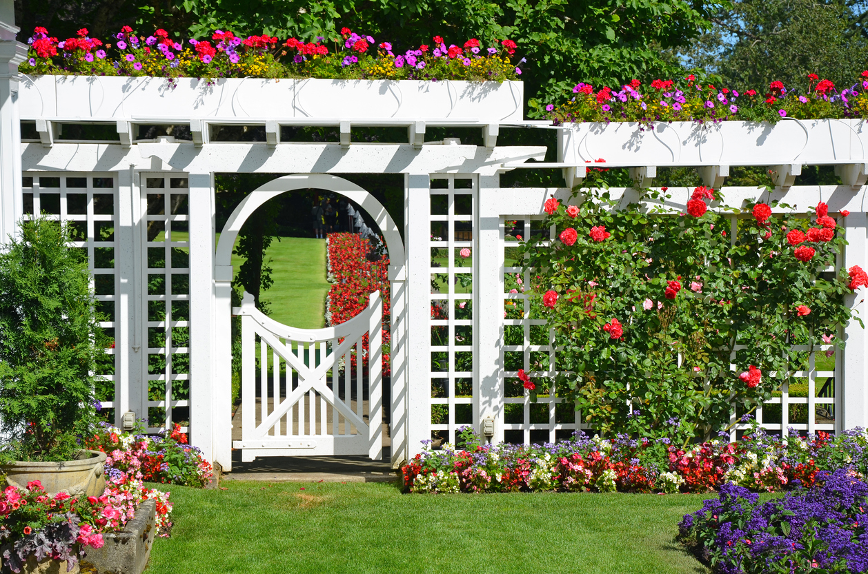 A large white ornamental garden gate and fence, surrounded by flowers. 