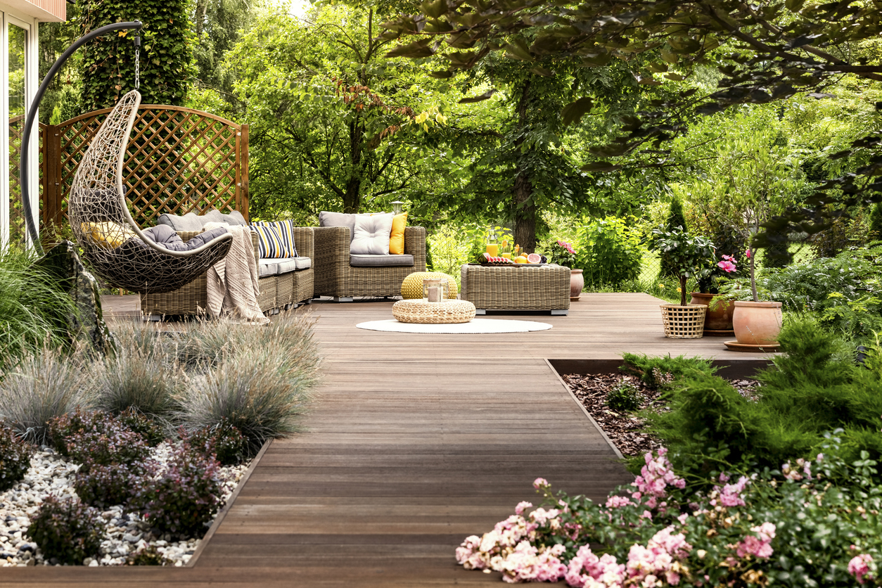 A wooden terrace surrounded by greenery, with wicker patio furniture and ornamental grasses.