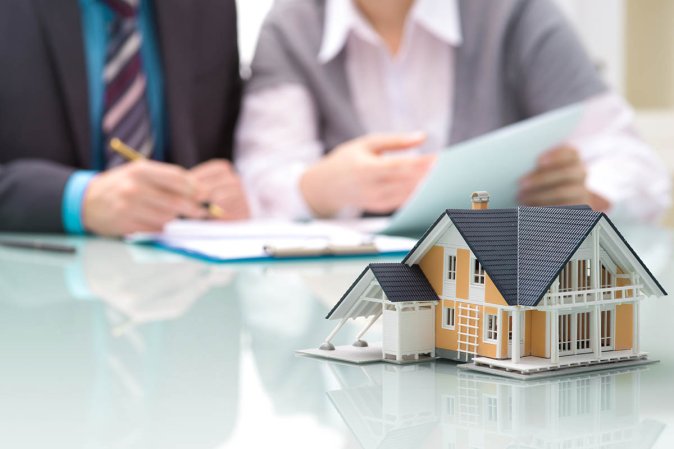 A glass table with a model house sitting atop, and people signing paperwork in the background.