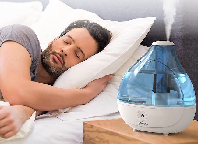 Man sleeps in a bed with an ultrasonic humidifier running on the nightstand next to the bed.