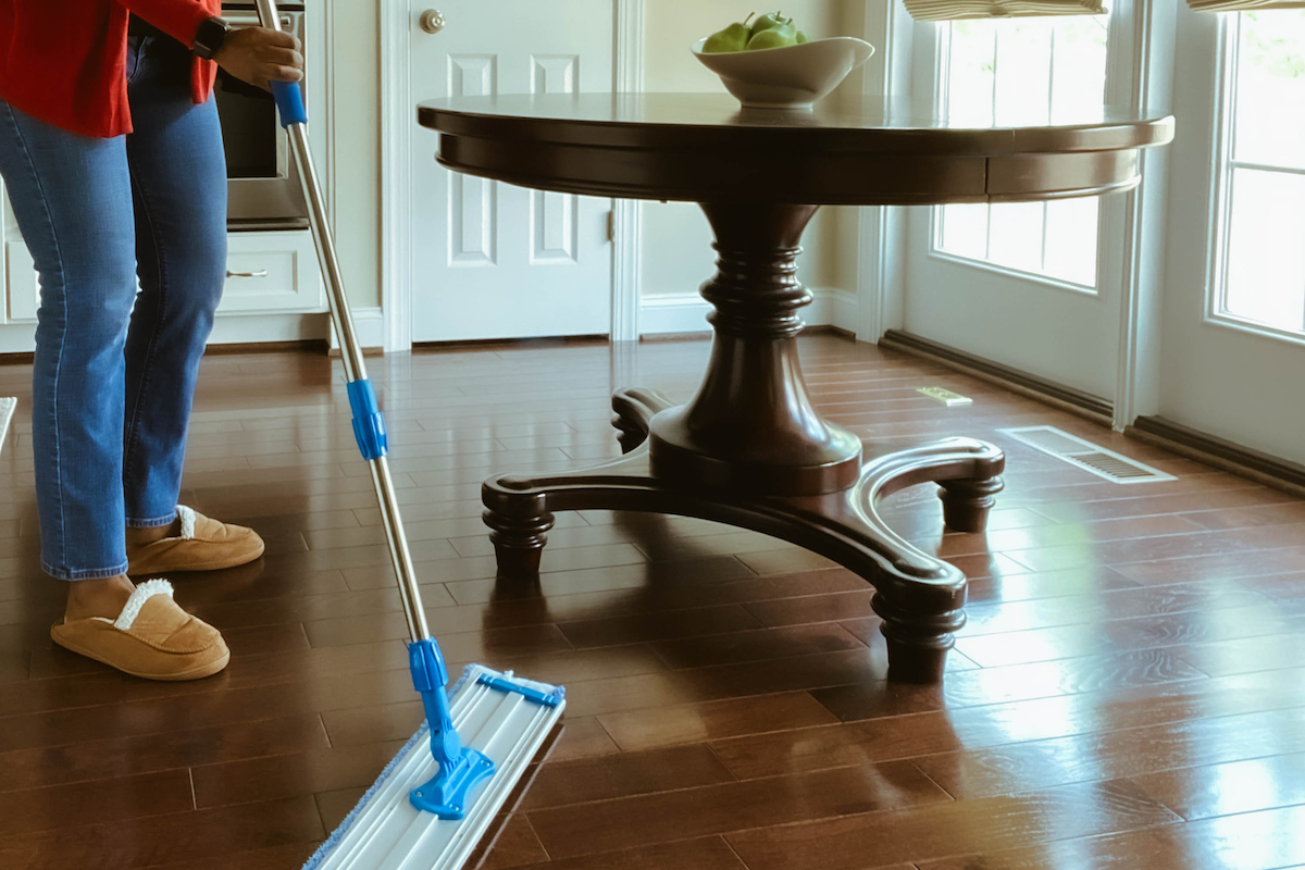 A person is using a dry mop to clean the hardwood floor around a wooden table.