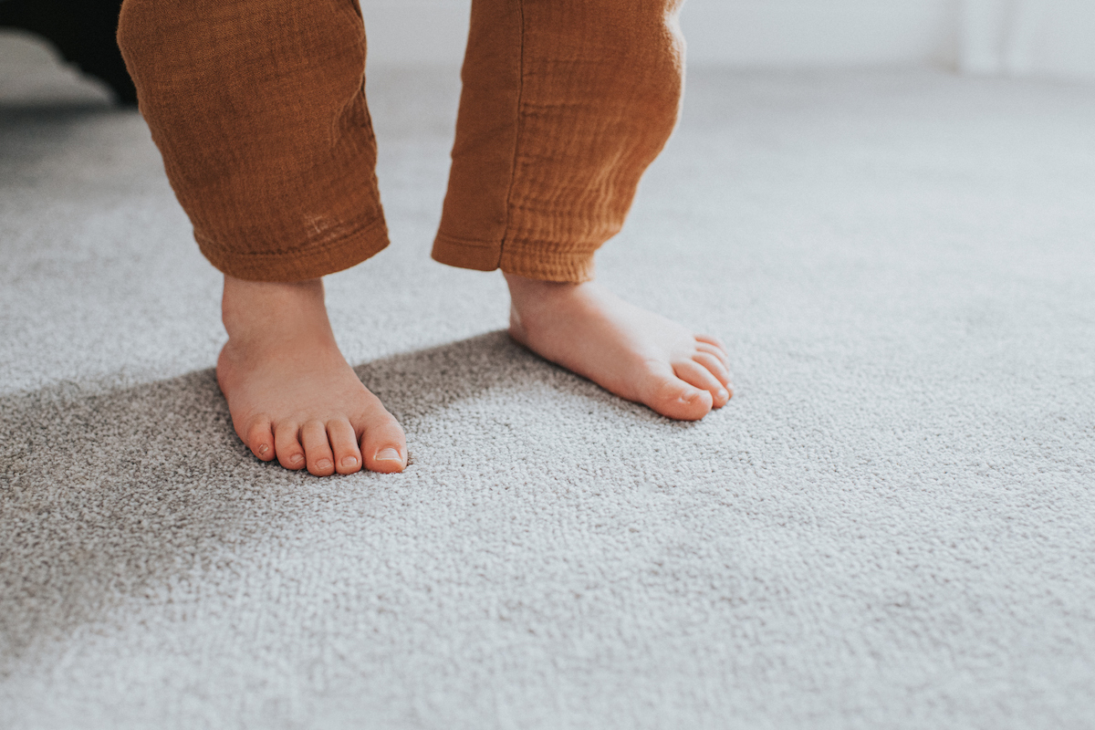 A young child stands on light grey carpet in the sunlight.