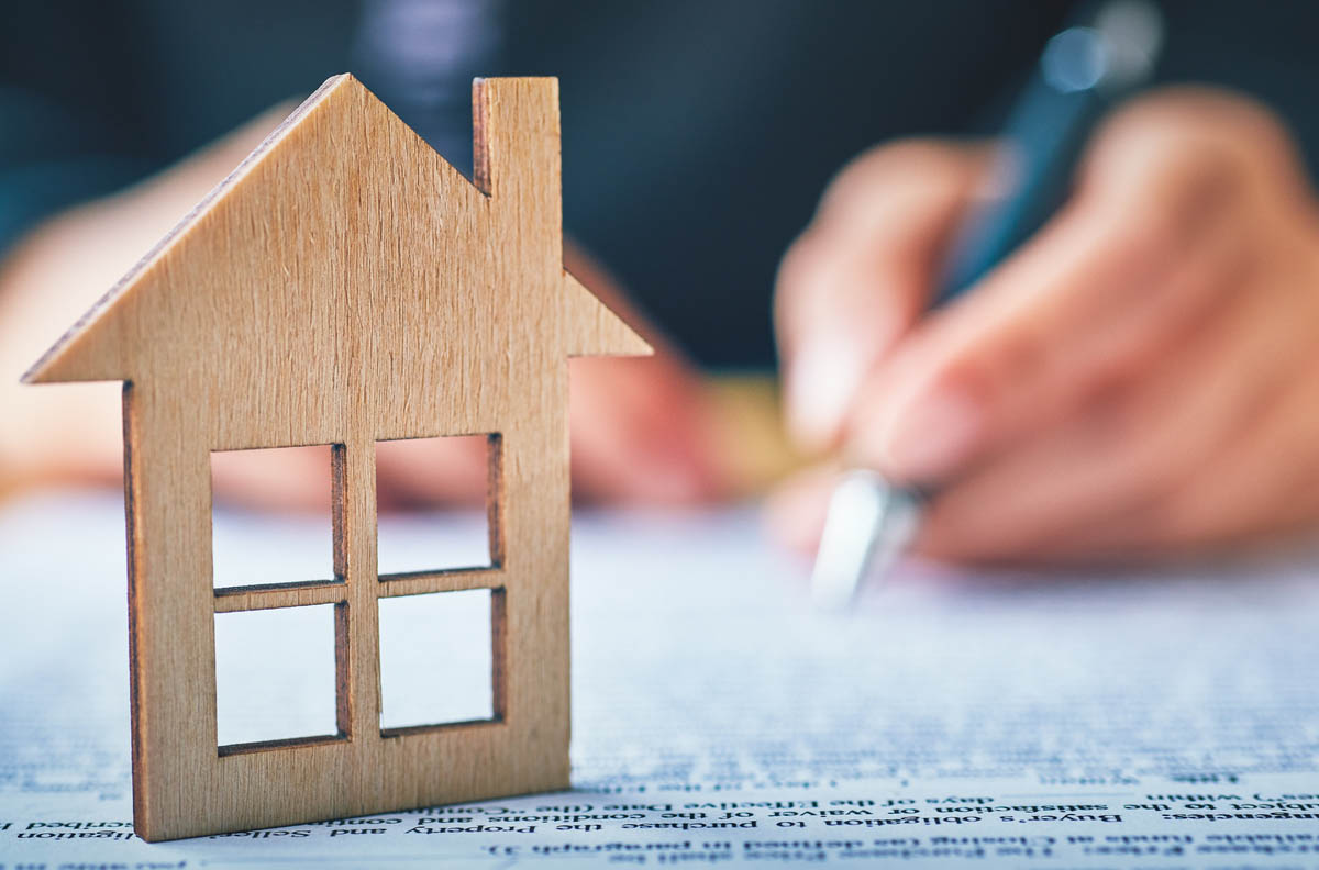 A small wooden model house facade sits on a table with a person writing in the background.