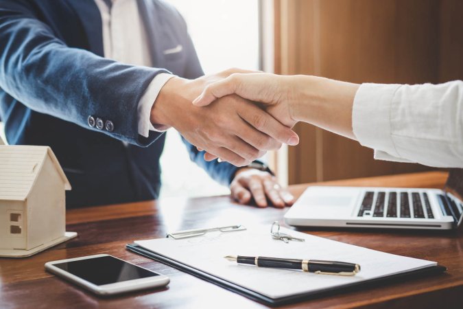 Two people shake hands at an office desk where a laptop, a phone, and a document sit.