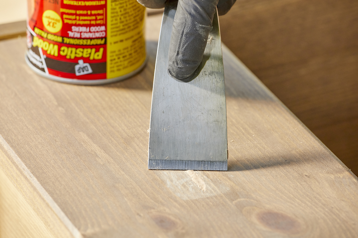 Woman uses a putty knife to apply wood filler to a nick in a wood board.