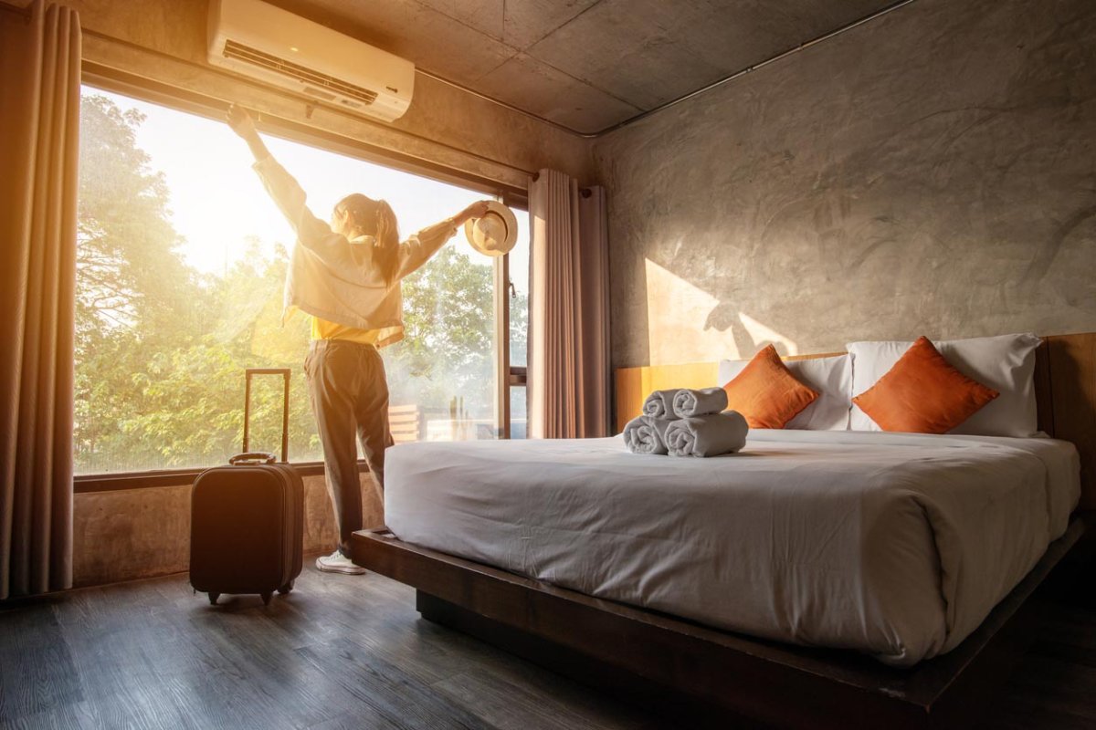 A person stands next to a bed, in front of a window with morning sun streaming in, and stretches.