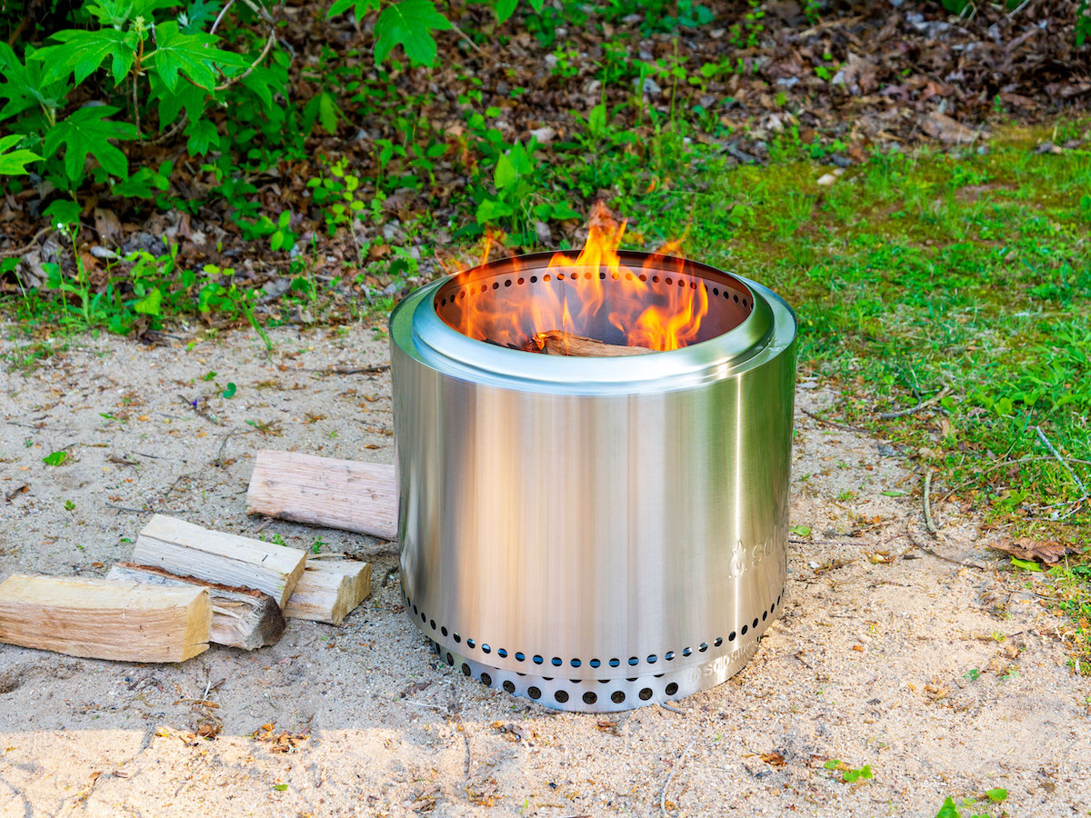 A silver metal smokeless fire pit has a fire burning kindling inside in a backyard.