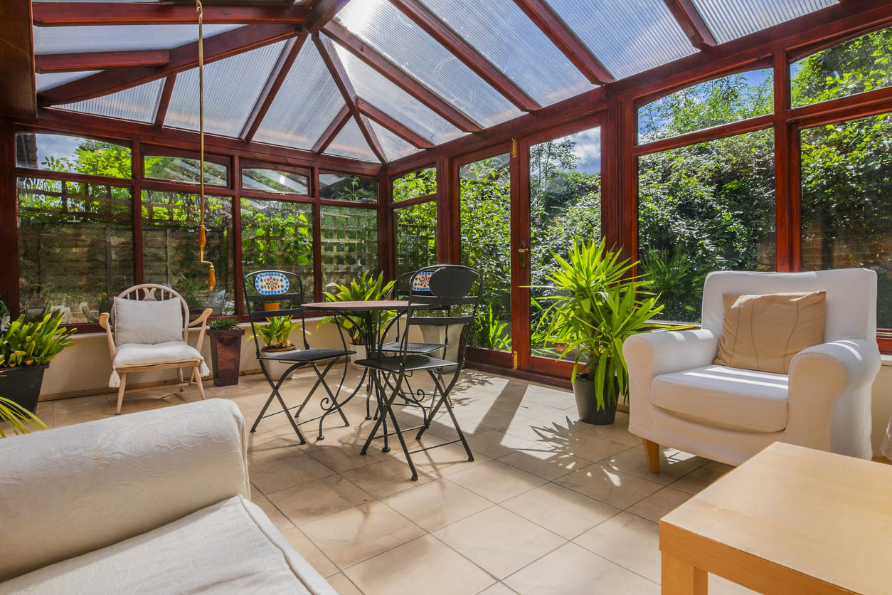 An interior shot of a decorated sunroom with glass walls and ceiling and wooden window frames.