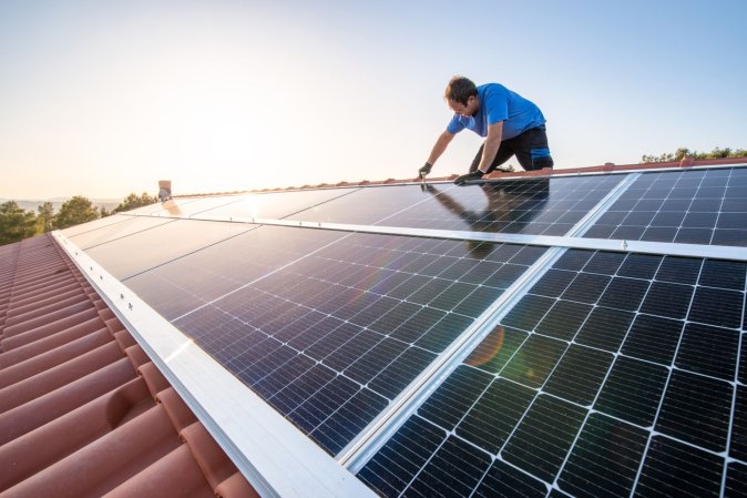 A view of a worker installing solar panels on a red tiled roof.