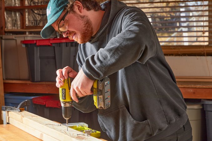 Man wearing a cap drills a screw into a piece of wood.