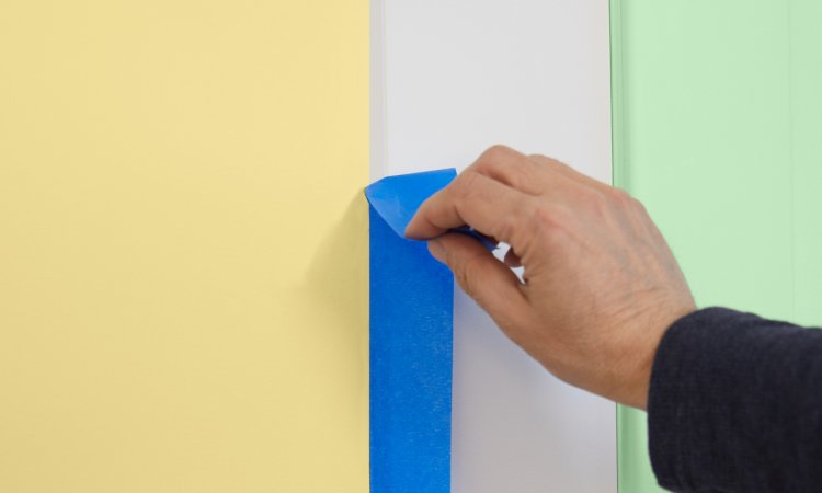 Person pulling painter's tape off white trim, with a yellow wall on one side and a mint green wall on the other.