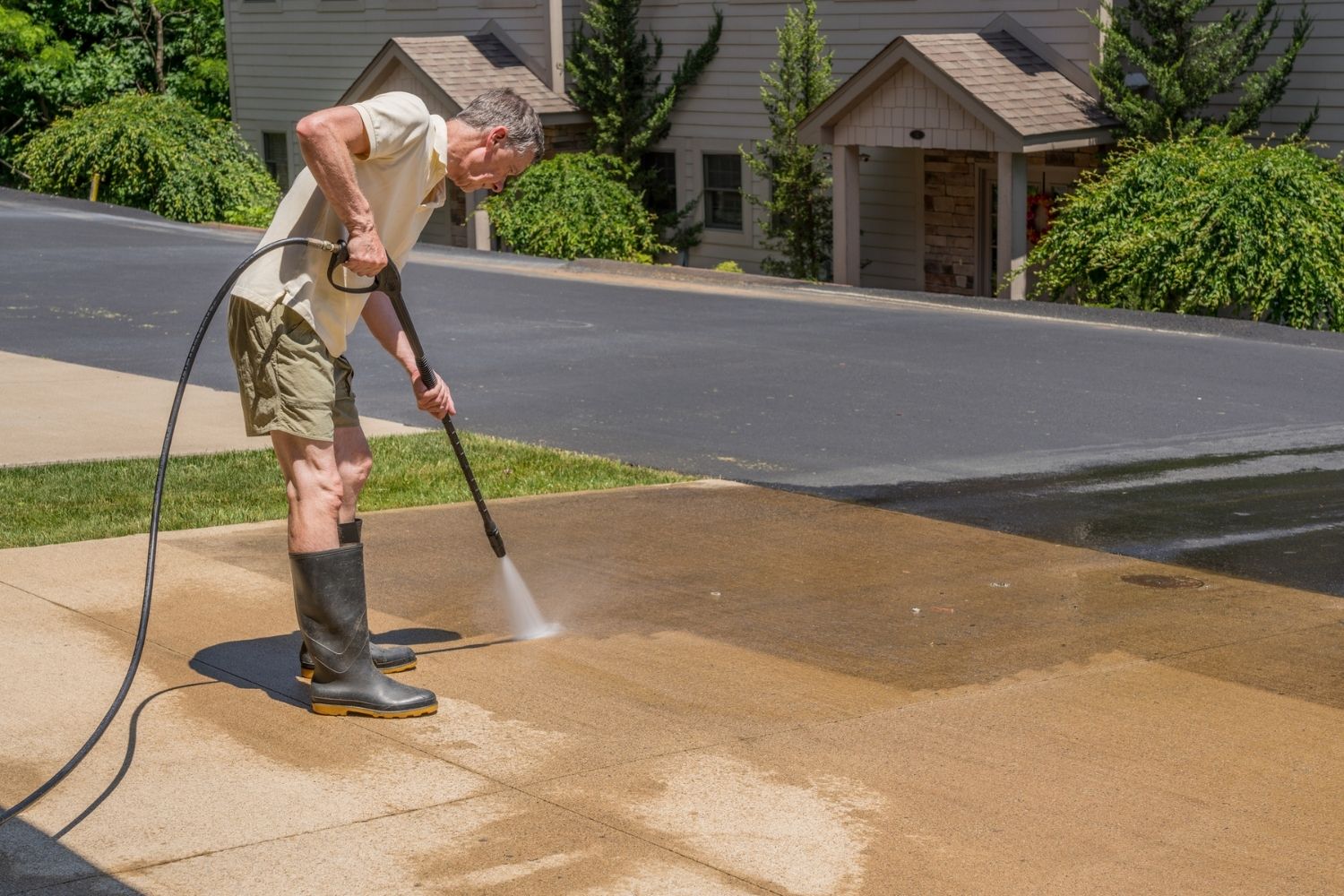 A worker wearing rubber boots pressure washes a concrete driveway in a residential neighborhood.