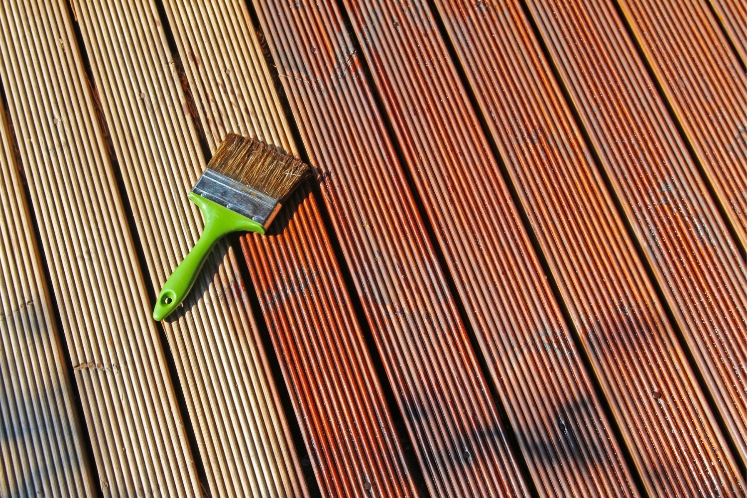 A close-up of a half-painted wooden deck with a paintbrush lying on top.