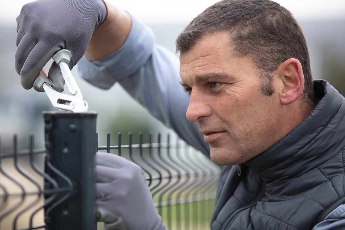 A worker wears gloves and uses pliers to install a fence.