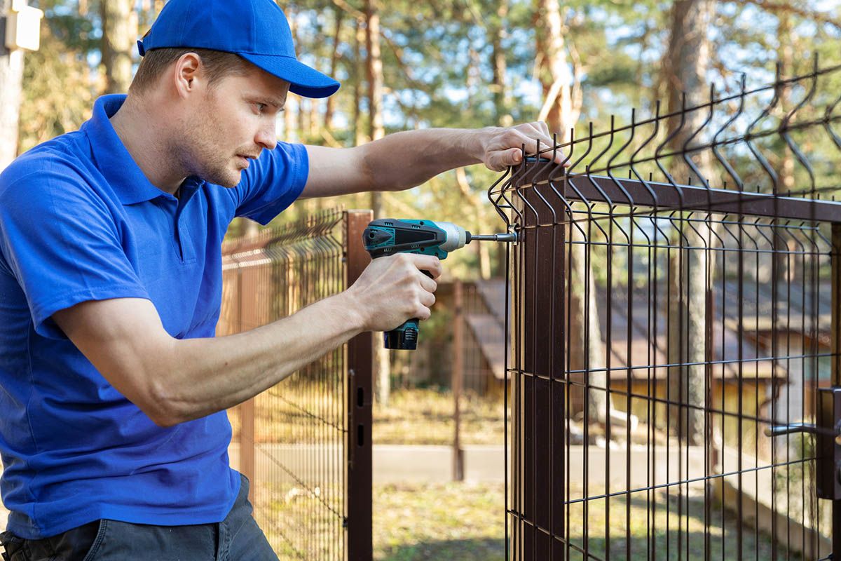 A worker in a bright blue uniform installs a fence using a drill.