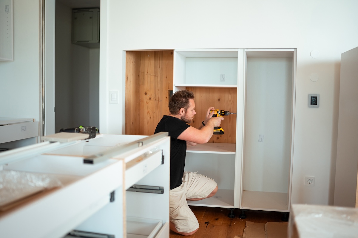 A man in a black shirt and khaki pants assembles white kitchen cabinets.