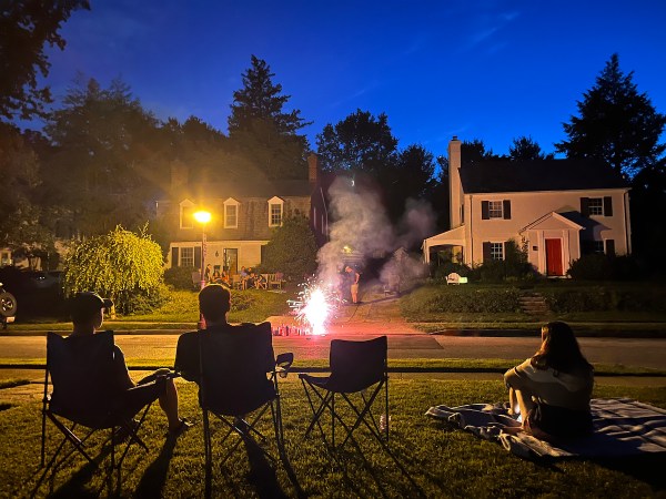 Neighbors in yard chairs watch fireworks exploding on street, with houses in background.