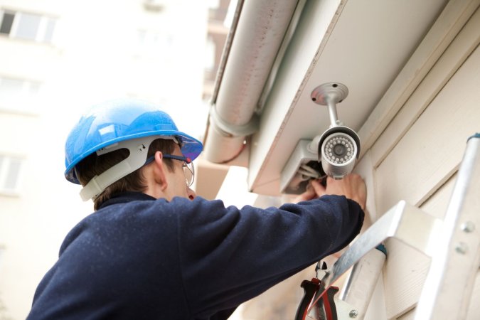 A person in a blue hard hat installs a security camera on the exterior of a home.