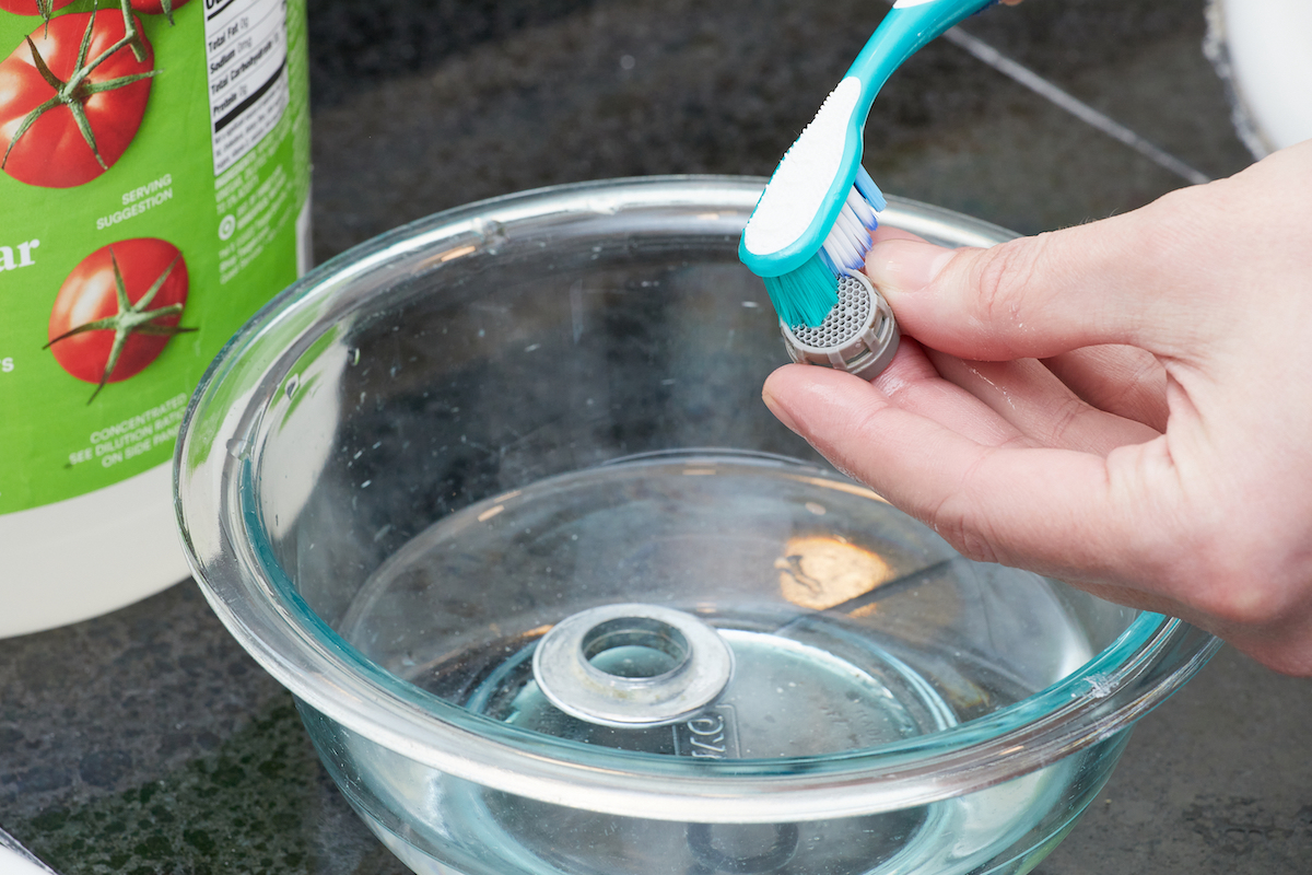 Woman uses a toothbrush to clean pieces of faucet aerator soaking in vinegar.