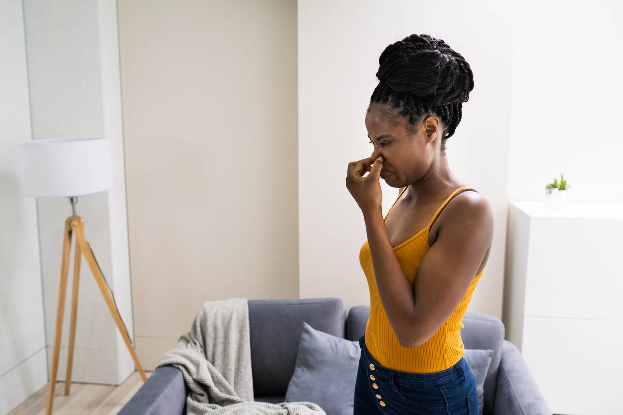 A person wearing a yellow top and blue pants stands in a living room and holds their nose as if there is an unpleasant smell.