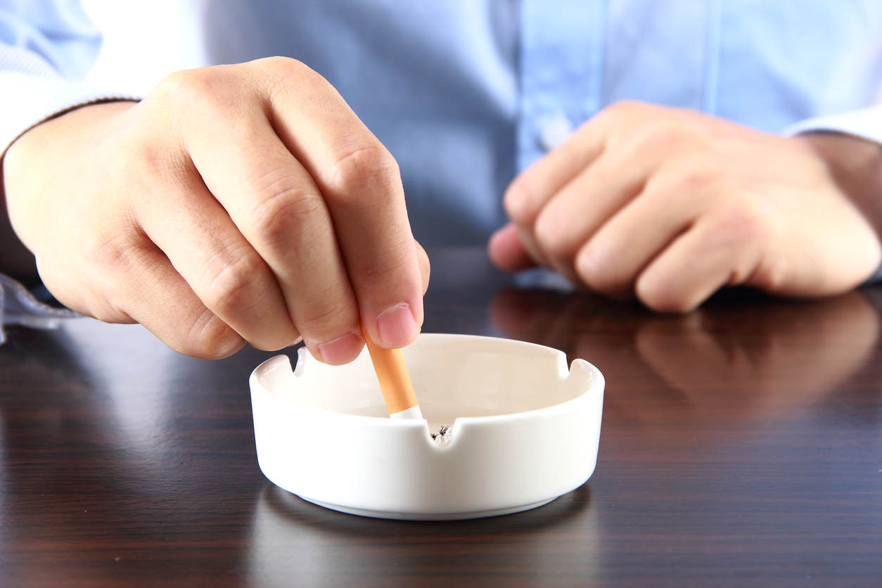 A close-up of a person stubbing out a cigarette in an ashtray atop a wooden table.