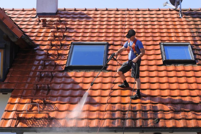 A worker is standing on a roof and spraying it clean with a pressure washer.