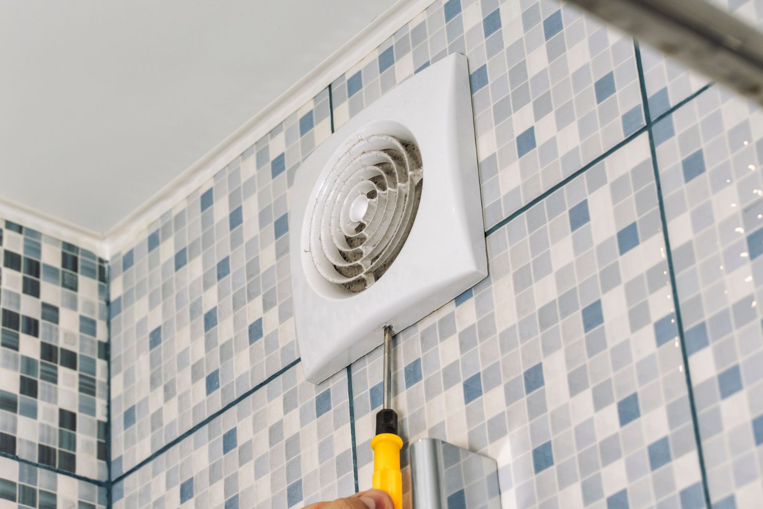 A person with only a portion of their hand visible uses a screwdriver to either remove or install an exhaust fan on a tiled bathroom wall.