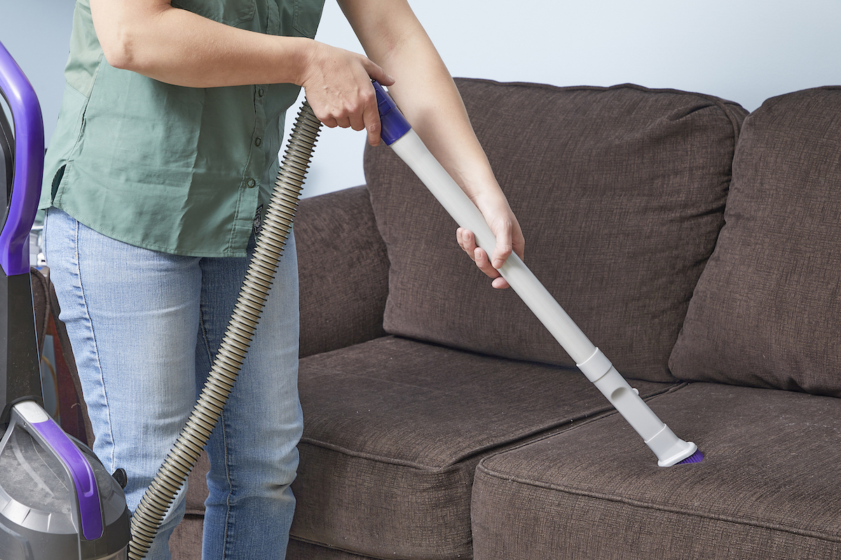 Woman uses an upholstery attachment to vacuum a brown couch.