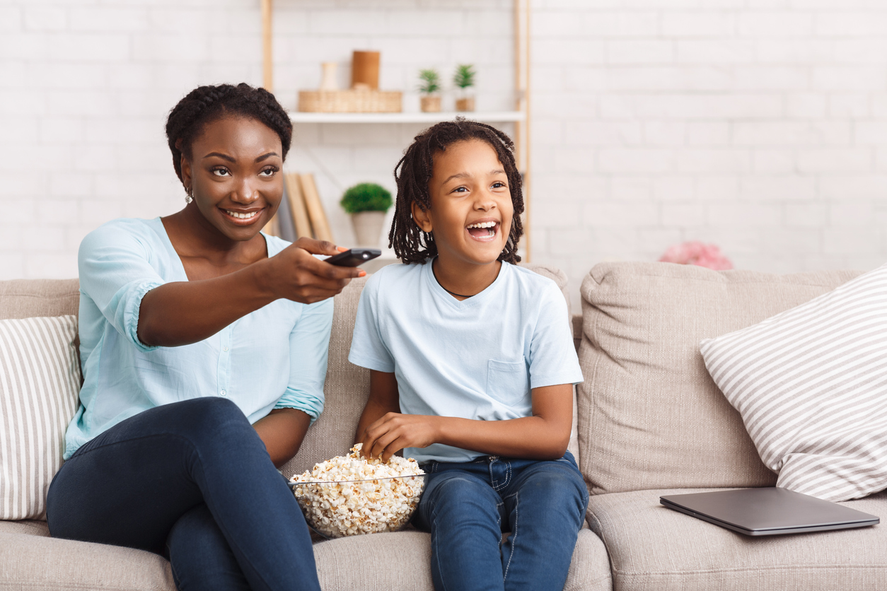 Woman and child sit on a couch with a remote control, eating popcorn.