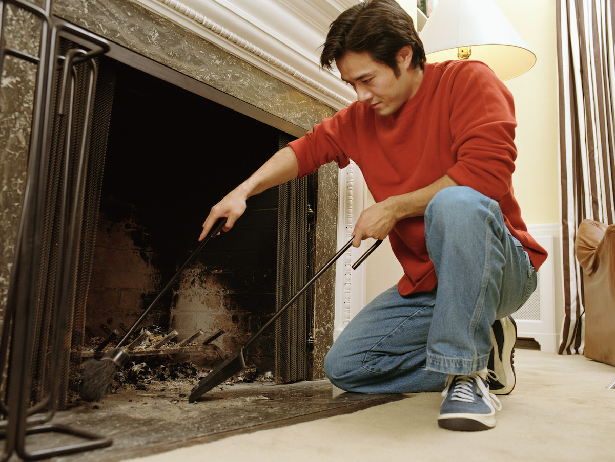 Man wearing a red sweatshirt uses fireplace tools to clean ash out of the firebox.