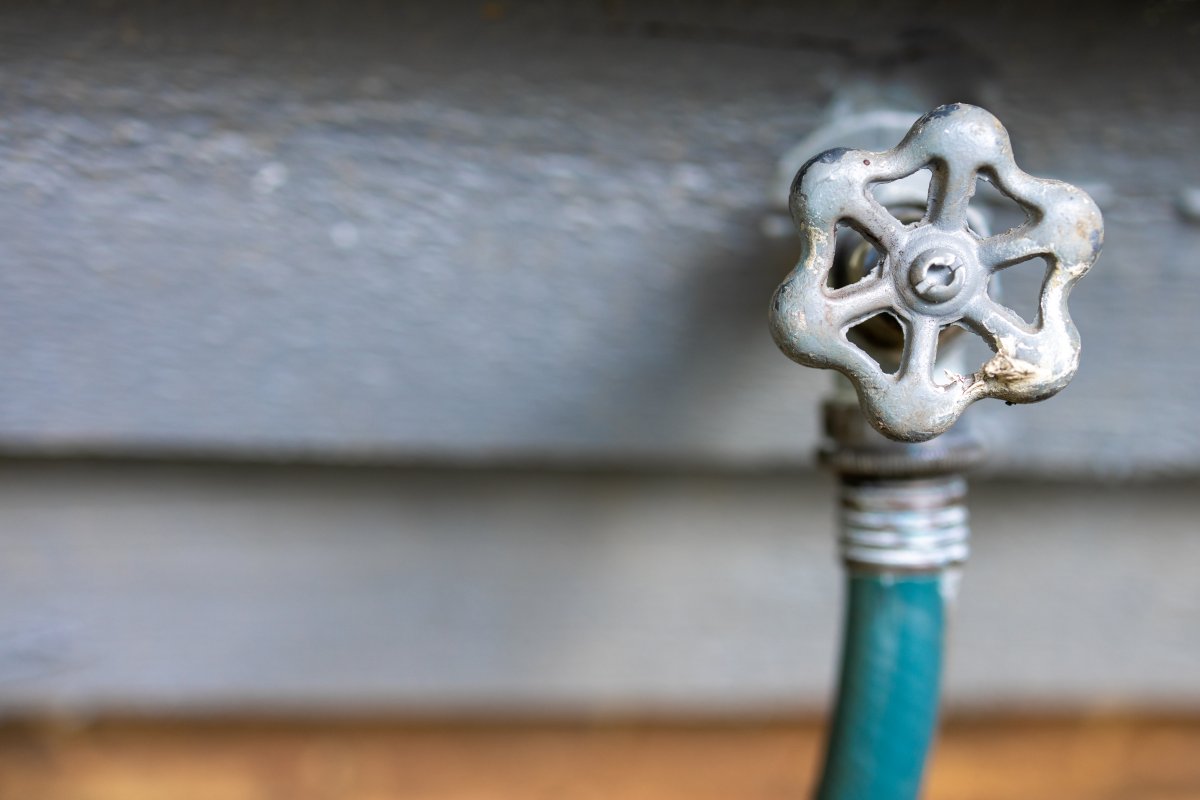 Outdoor water spigot connected to a green garden hose, grey vinyl siding in the background.