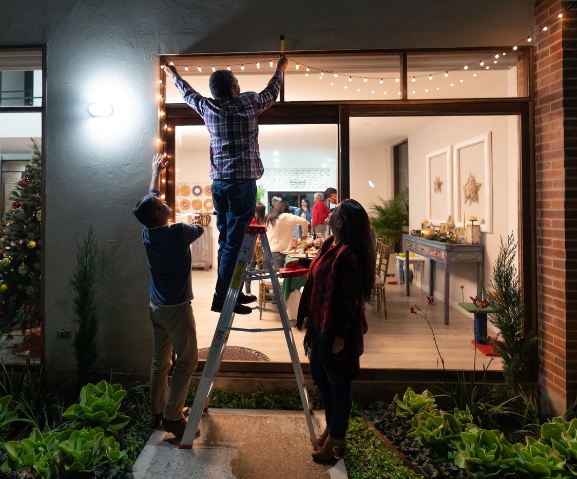 Friends hanging outdoor string lights on a patio, lights indoors illuminated.