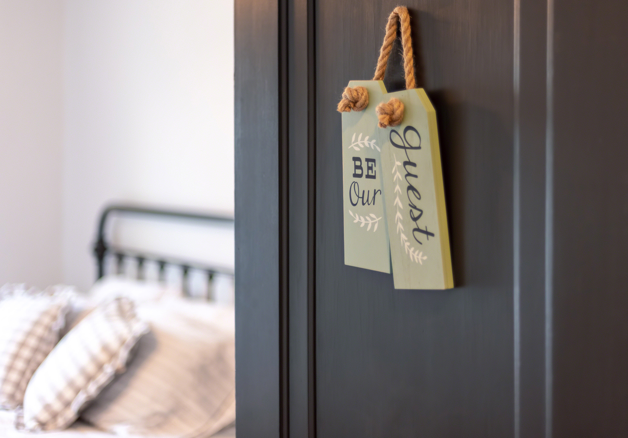 A guest bedroom door ajar, with a bed inside room and a "Be our guest" sign on the door.