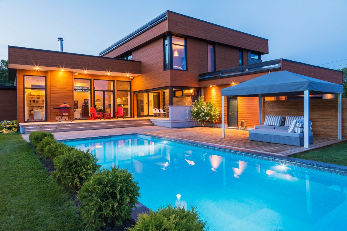 A modern,, rectangular home with a lit swimming pool that sits aside deck awning.