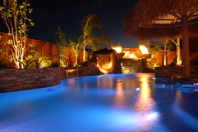 Swimming pool at night lit with in-water blue lights and warm light fixtures surrounding a fountain.