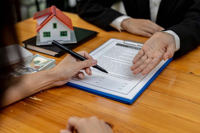 A person signs a document on a clipboard as a person in front of them gestures.