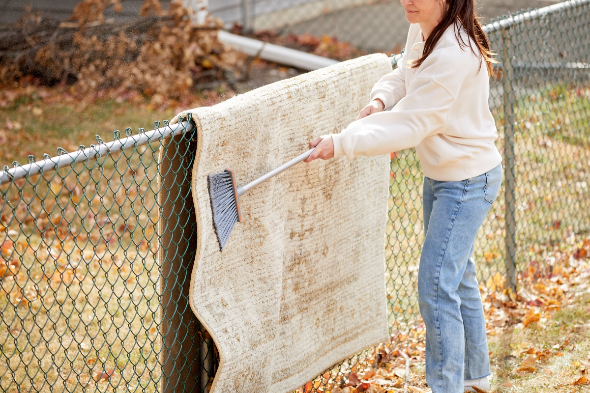 A woman is using a broom to beat dust out of a carpet that's hung on the fence.