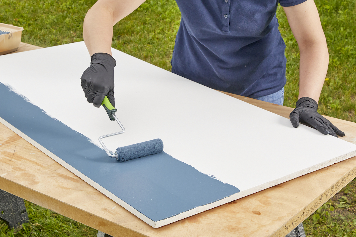 Woman uses a small paint roller to apply blue paint to a piece of particle board.