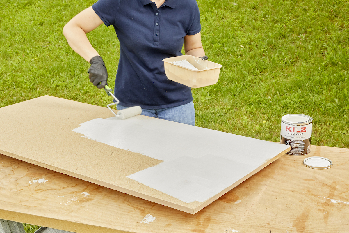 Woman primes a large piece of particle board on a table outdoors.